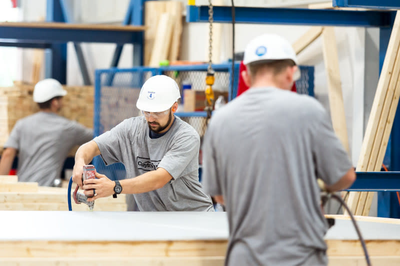 Three men in gray T-shirts and white hard hats, two of them with their backs to the camera, work with drills on a manufactured home inside of a home building facility.