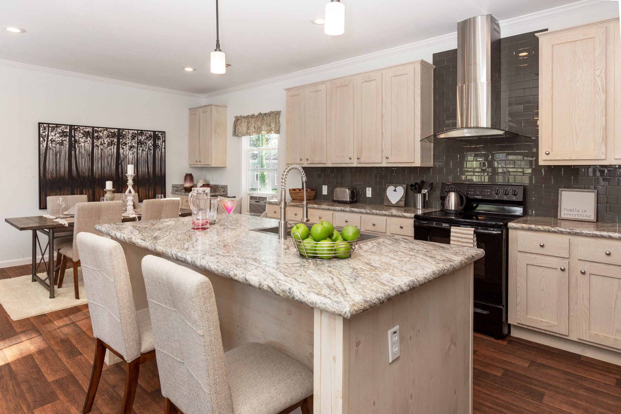 Manufactured home kitchen features a large island with a beige marbled countertop and also dark, grey glass backsplash with a silver hood over it.