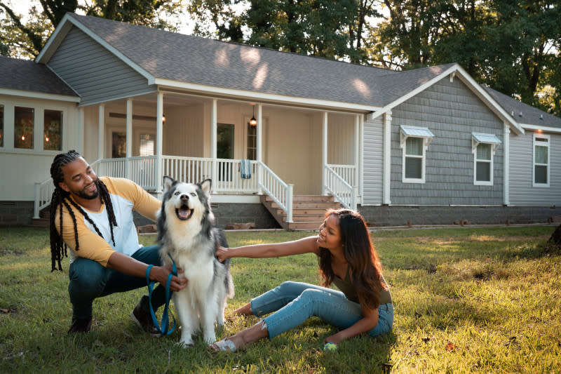Man and woman pet dog while sitting on the lawn in front of a manufactured home with a gray roof, gray siding and a wraparound porch with white railings.