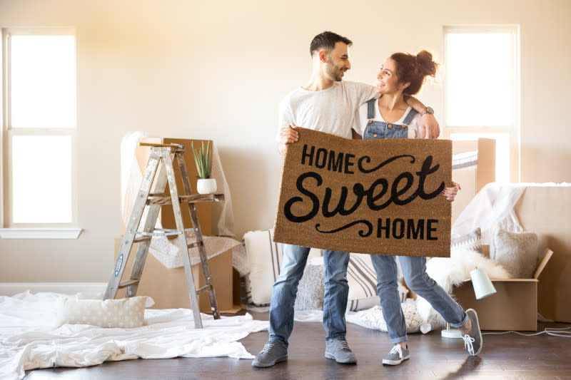 Young couple in jeans and white shirts holding welcome mat that says “Home Sweet Home” with boxes, ladder and sheets and pillows on the floor behind them.