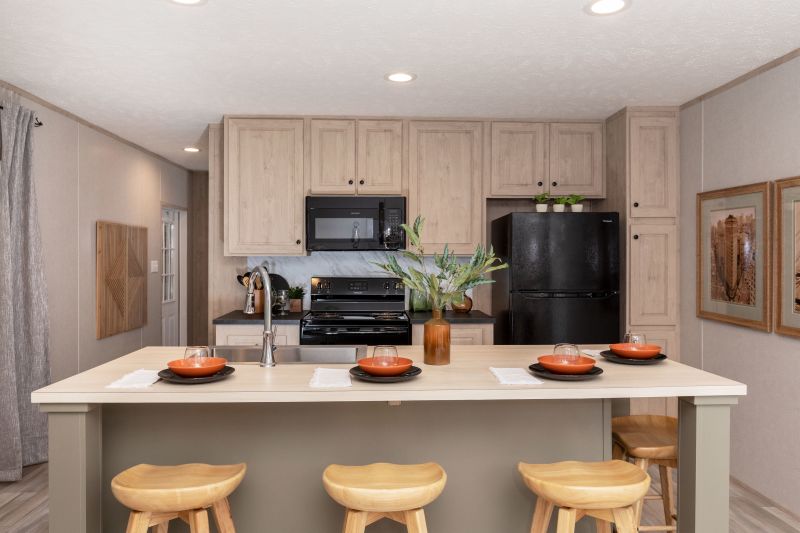 A manufactured home kitchen is in view with a large island/ breakfast bar, the cabinets a light brown, and black appliances.