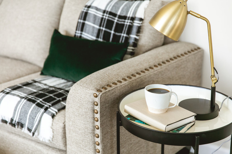Picture of a tan couch with a plaid blanket and green throw pillow, with a gold lamp on a table next to it.