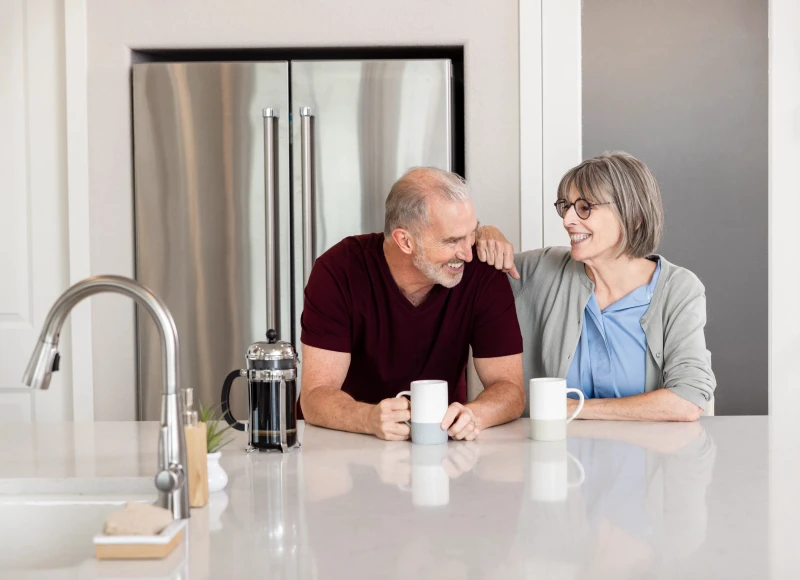 Man and woman smile while sitting at a kitchen island, with coffee cups in front of them and a fridge in the background