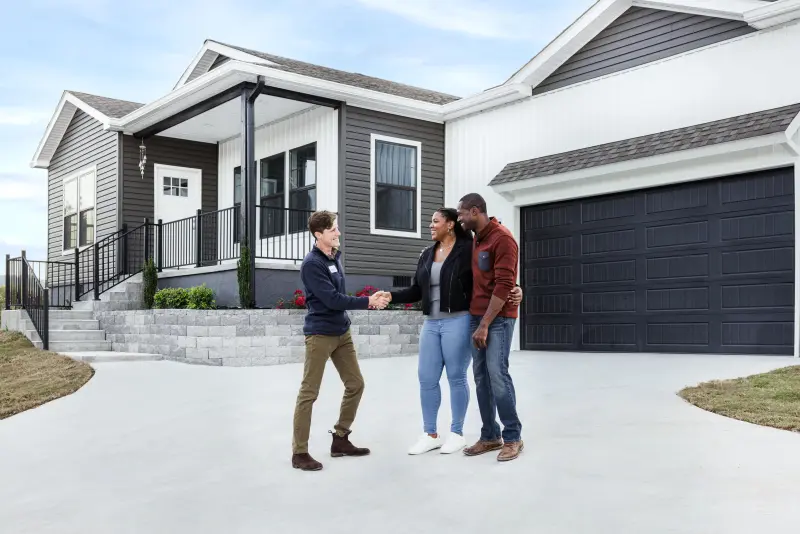 A couple stands in front of an attached garage of a manufactured home, shaking hands with a home center consultant.