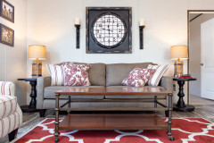The living room of a manufactured home with a gray couch with red and white pillows to match the rug and chair, a wood and metal coffee table, black end tables with gold lamps on top and a gray square clock hanging on the wall.