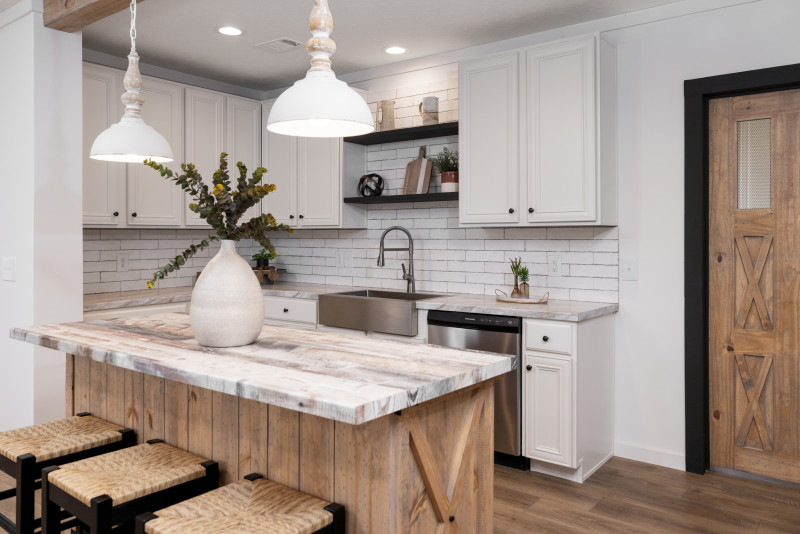 The kitchen is in view with white cabinetry and backsplash. There’s a wooden island with a white marble-style countertop, and farmhouse lights hung over it.