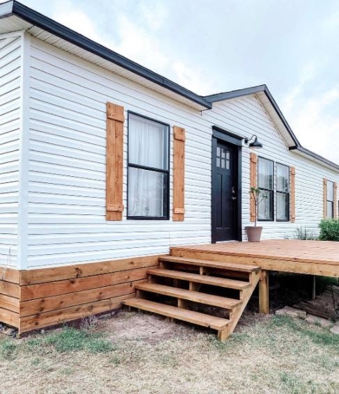 Manufactured home with white vinyl siding, cedar shutters, cedar skirting and black trim around windows and doors.