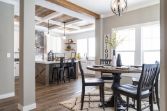 View is of the corner of a dining room with natural light flowing in. On the other side the kitchen can be seen with a tray wood ceiling with white beams and a kitchen island with chairs. 