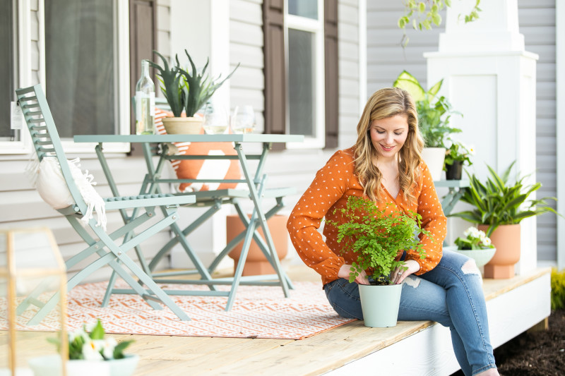 Woman in an orange shirt and jeans packs soil around a potted plant while sitting on the edge of a front porch of a manufactured home with patio furniture, planters and other plants in the background behind her.