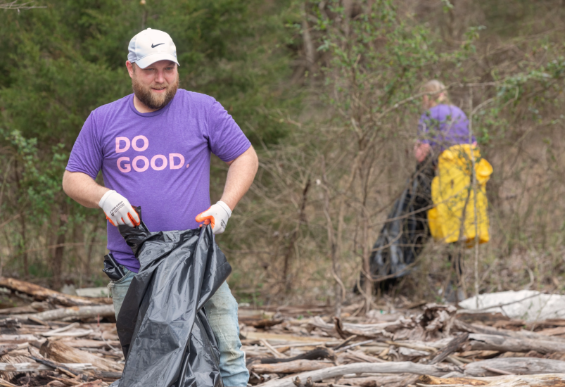 Man in purple "Do Good" shirt collects trash in bag along a river bank