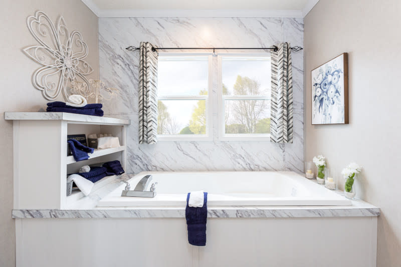 A bathtub in a manufactured home with built-in shelving and a white marble look accent wall.