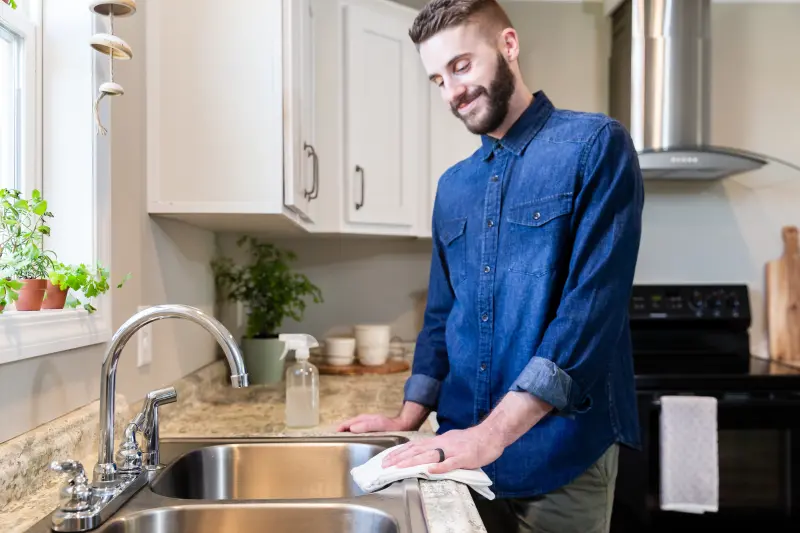 A man in a denim shirt wipes down the counter around a stainless steel kitchen sink.