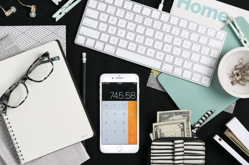 Flat lay of budgeting items and office supplies featuring a white keyboard, calculator, wallet with cash and a notebook with a pair of glasses on top.