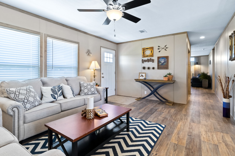 The living room in this manufactured home has off-white and grey couches with a brown coffee table in front of them. There’s a zig-zag white and black rug on the floor underneath the table. View shows a hallway into the rest of the home.
