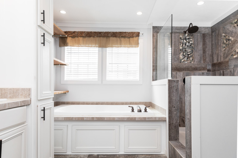 The view in this manufactured bathroom includes a white soaker tub, some cabinets with black door pulls, and a walk-in, partially open shower with a glass panel and brown stonework shower detail.