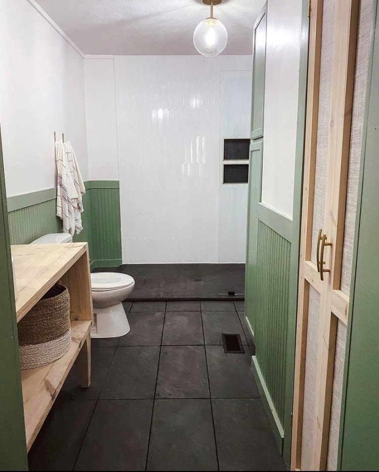 Manufactured home bathroom with black straight stack tile, large wood vanity, sit on top sink, green wainscoting, and a walk-in shower with white shower tile on the walls, black tile on the shower floor and gold faucets and rainfall showerhead.