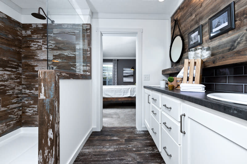 Manufactured home bathroom with wood-paneled doorless shower and vanity with white cabinets, dark countertop and dark title backsplash.