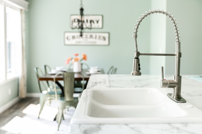 View of a white sink with stainless steel detachable sink nozzle. There is a pastel green dining room in the background with a set table.