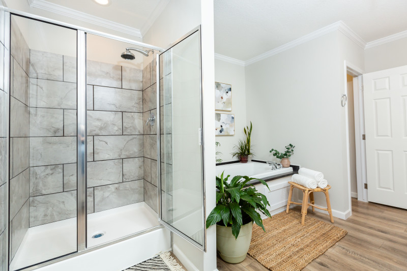 There is a walk-in shower with a gray tiled wall and the door hinged open. There’s a bathtub set back with green plants around it, a brown floormat, and a roll of towels.