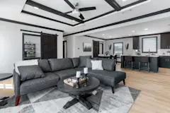 A sliding barn door is open to a room behind a modern gray lounge sofa and round coffee table in the living room that connects to a kitchen with black cabinets and an island.