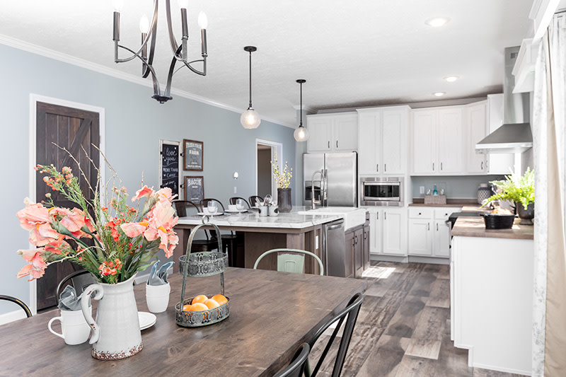 Dining area and kitchen of the Country Aire manufactured home with wooden table, metal chairs, pendant lights, wooden island and white cabinets.