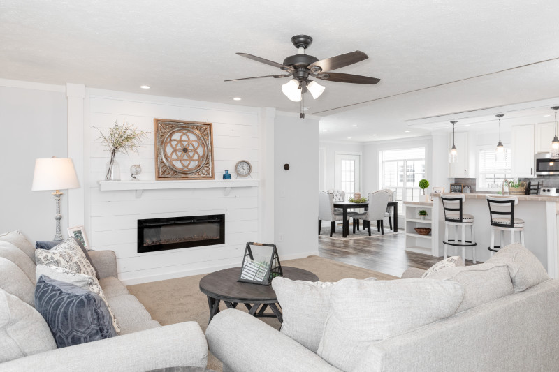 The living room is white with a large fireplace and modern design. There is a brown ceiling fan, brown table, and two white couches. The kitchen and the dining area are also in view in the distance.