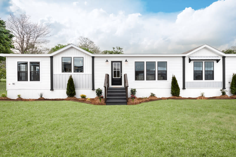 A white manufactured home sits on a landscaped yard with a mulched flower bed and small trees and has vinyl siding, two dormers on either end, a window awning and a black front door and hardware lights.