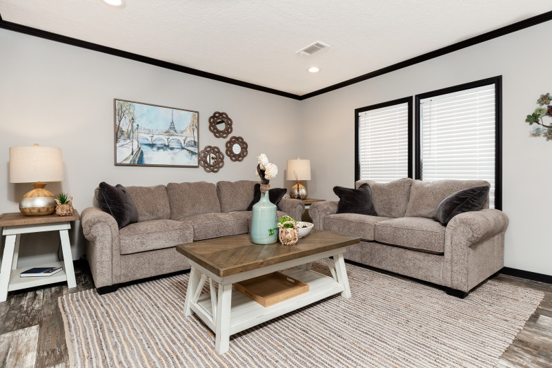 Living room of a manufactured home with dark trim and gray wood floors, neutral furniture.