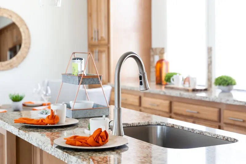 Stainless steel Pfister faucet in the kitchen island of a manufactured home, with brown granite countertops, tan cabinets and cream, orange and green decor.