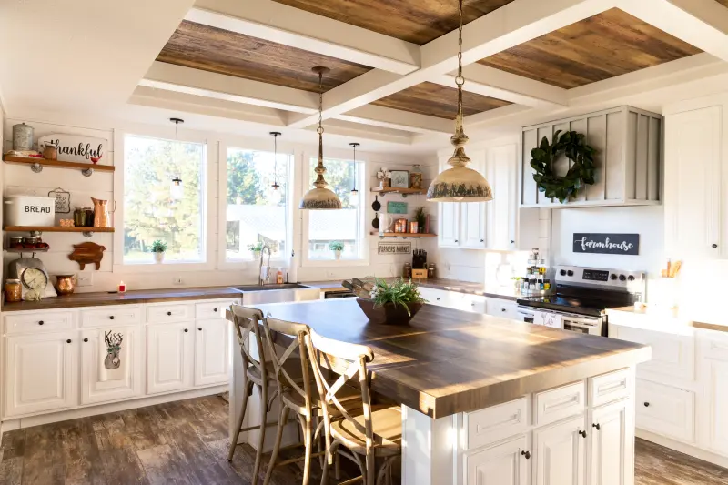 A farmhouse style kitchen in a manufactured home with a large kitchen island, rustic ceiling beams, open shelving and antique style pendant lighting.