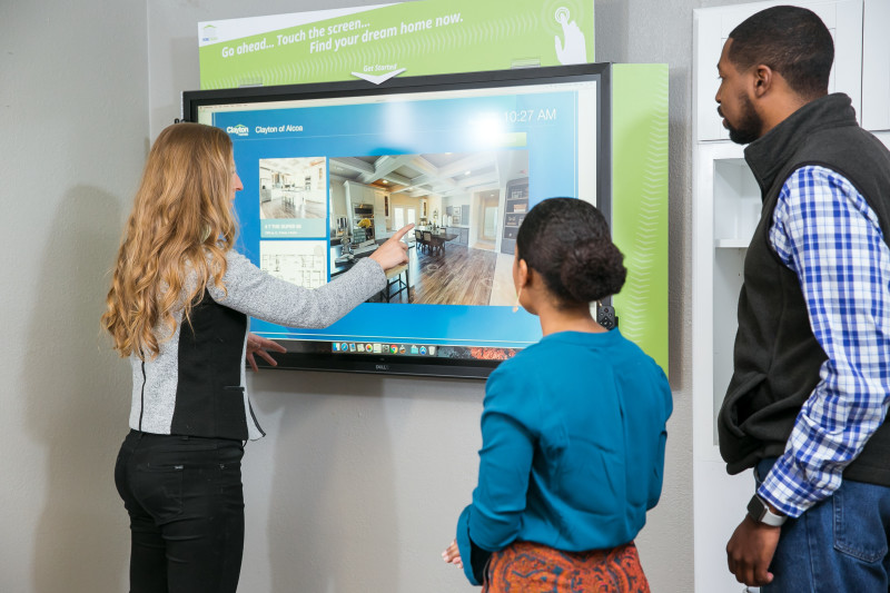 Woman points and shows a man and woman a Clayton home floor plan on a touch screen inside of a Clayton home center.