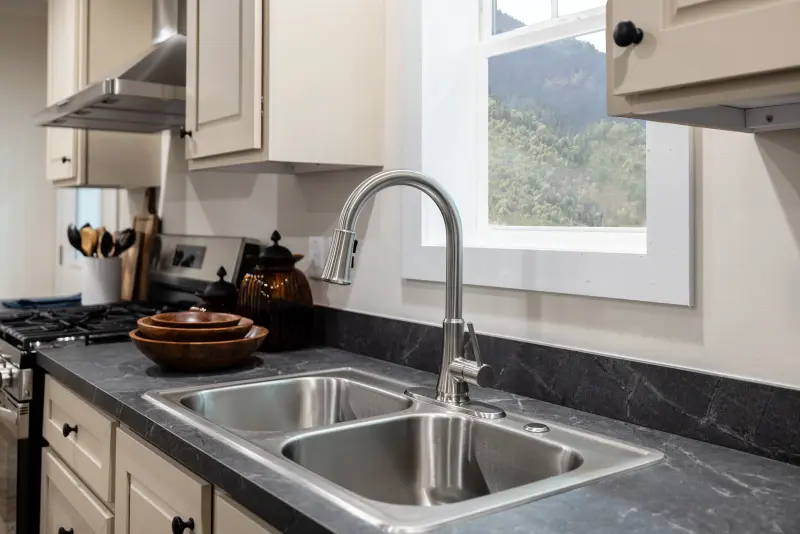 Close-up of a stainless steel sink and faucet in the kitchen of a Clayton manufactured home with light tan cabinets and dark marble countertops.