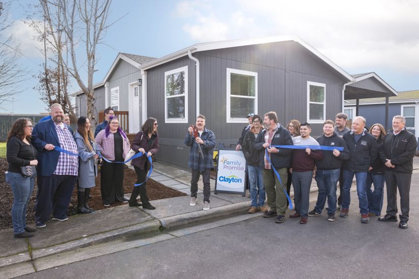 Man cuts blue ribbon in front of sign that reads "Family Promise + Clayton" and an off-site built home