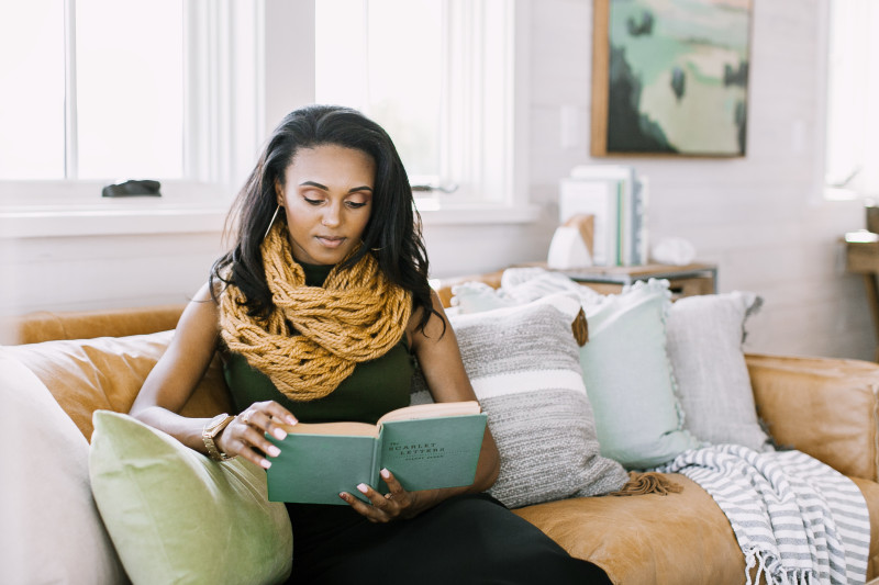 A woman reads a blue book on a tan leather couch with throw pillows.