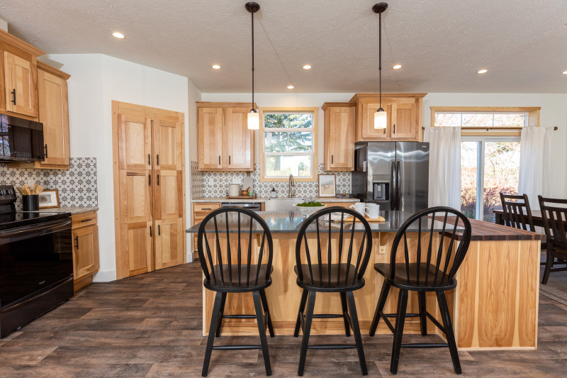 Image is of a manufactured home kitchen. The theme of the cabinets is a yellowish wood with black bar pulls. There’s a huge island with three black chairs pulled up to it. There’s tons of lighting and cabinets as well.