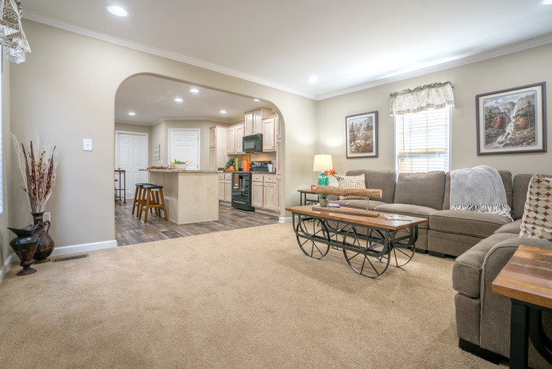 Manufactured home living room with carpet and a large entryway into the kitchen.