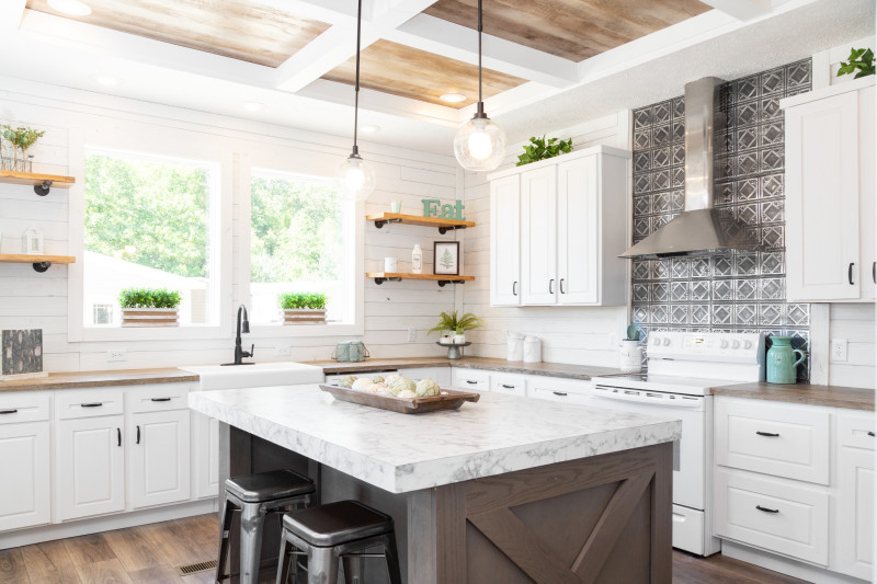 Farmhouse kitchen of a Clayton manufactured home with white cabinets, white shiplap walls, a tray ceiling, a kitchen island and two large windows.