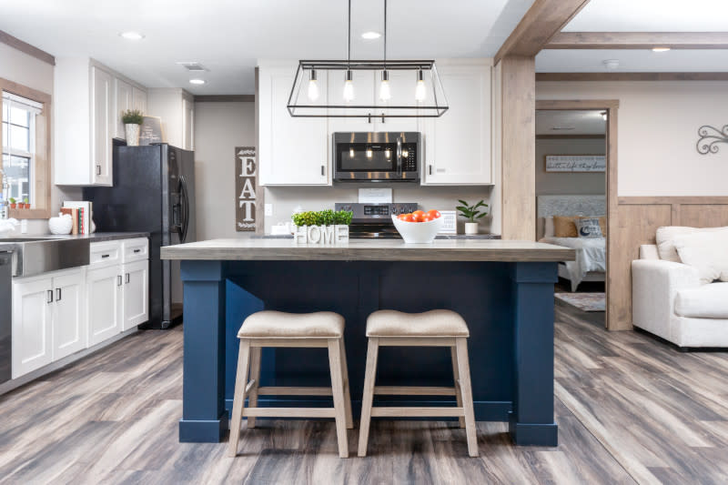 Manufactured home kitchen with white cabinets, stainless steel appliances and blue kitchen island with 2 stools, with the edge of the living room and the primary retreat visible on the right.