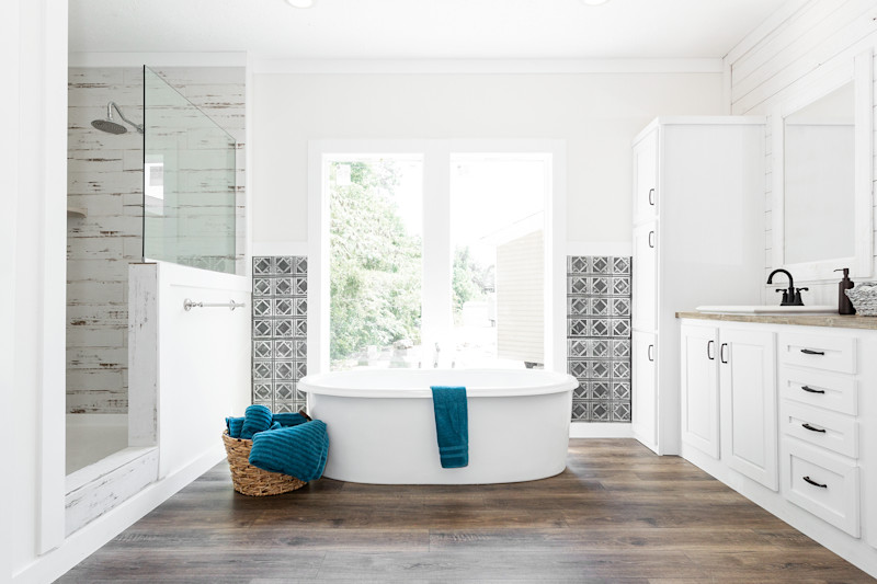 Primary bathroom in a Clayton manufactured home with a glass wall shower, vanity with white cabinets and oval bathtub in front of a window.