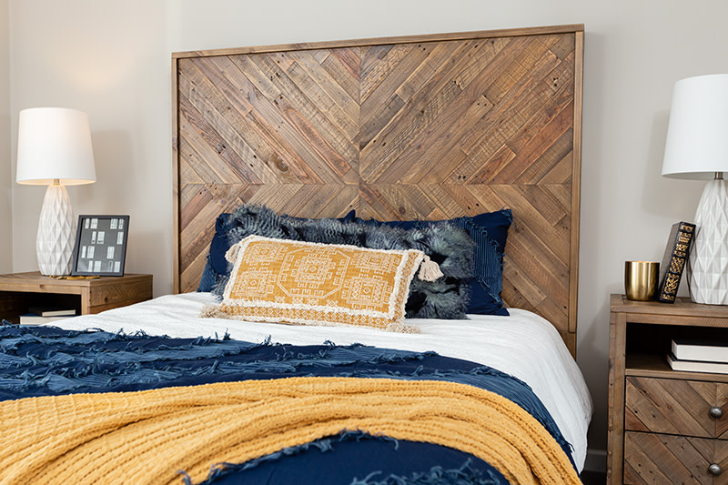 Bedroom with reclaimed wood headboard and yellow, navy and white bedding, with 2 matching wood bedside tables with white lamps and books.