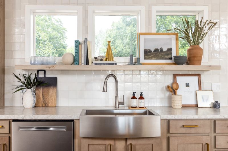 There are three square windows above a shelf in a modern, white tiled kitchen. Stainless steel sink and white countertop sit below the windows in this kitchen.