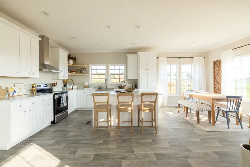 A kitchen with all white cabinets and counters and a large kitchen island that opens up into a dining area with large windows and a long wooden table with benches and chairs for seating.