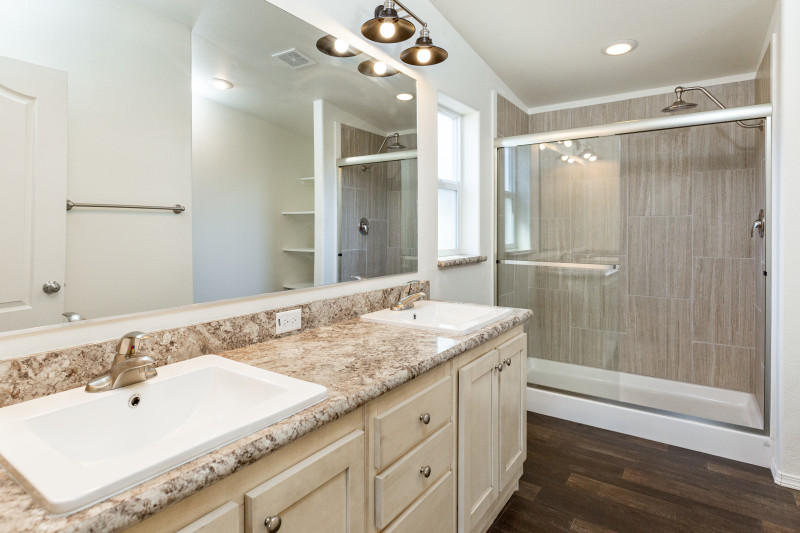 This luxury bath has a double vanity with crème-colored cabinets. To the back of bathroom there’s a walk-in shower with sliding glass doors. The bathroom floor is a dark colored wood.