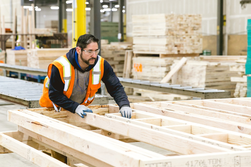 Man wearing a safety vest and glasses builds part of a prefabricated home inside of a facility, with more lumber in the background