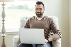 Man in a brown sweater and glasses looks at a laptop screen while sitting in a chair, with a window and lamp behind him.