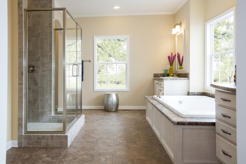 There's a side view of a large bathroom with a walk-in, glass door shower, a soaker tub, and vanities on either side of it. The walls are a light yellow and the floor and bathroom tile accents are brown.