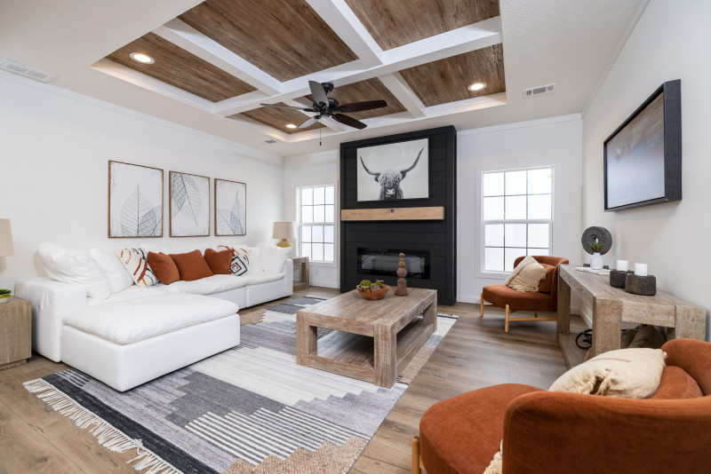 A manufactured home living room with wooden tray ceiling in a white room. There’s a dark gray, modern fireplace on one wall with windows on either side of it. Furniture is white and orange.
