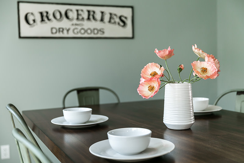 Dark wood table and green metal chairs with 3 white plates and bowls and a white vase with pink flowers, with green walls behind it and a sign that reads “Groceries and Dry Goods.”