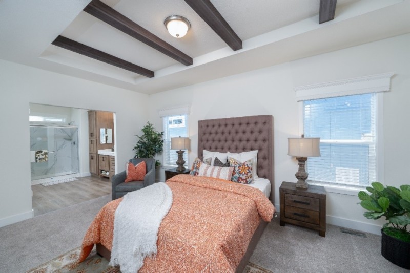 Bedroom in the Coronado 3760A with tray ceiling, wooden beams, bed with headboard, bedside tables and a chair. The room is white with carpet and connects to the bathroom.