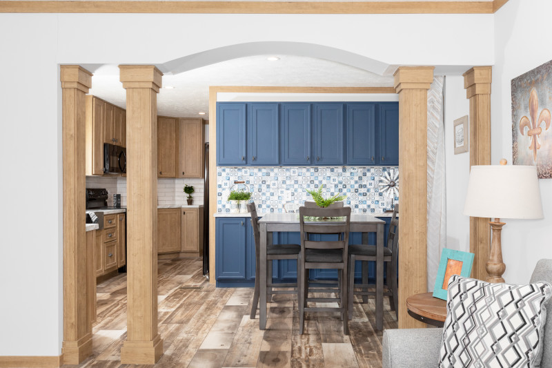 Dining area and kitchen of a single wide manufactured home with light wood columns and trim in an archway leading to blue accent cabinets, patterned tile backsplash and wooden table and chairs.